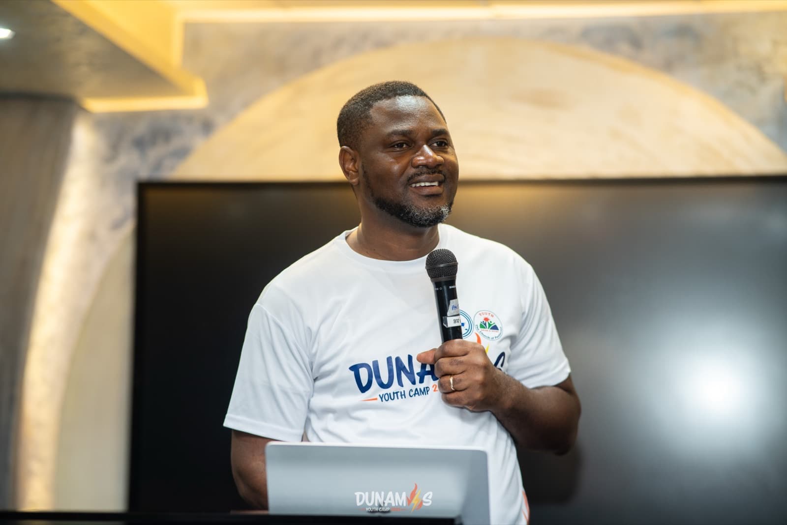 Elder Nana Sakyi Baffoe speaking into a microphone at Dunamis Youth Camp 2026, wearing a camp T-shirt.